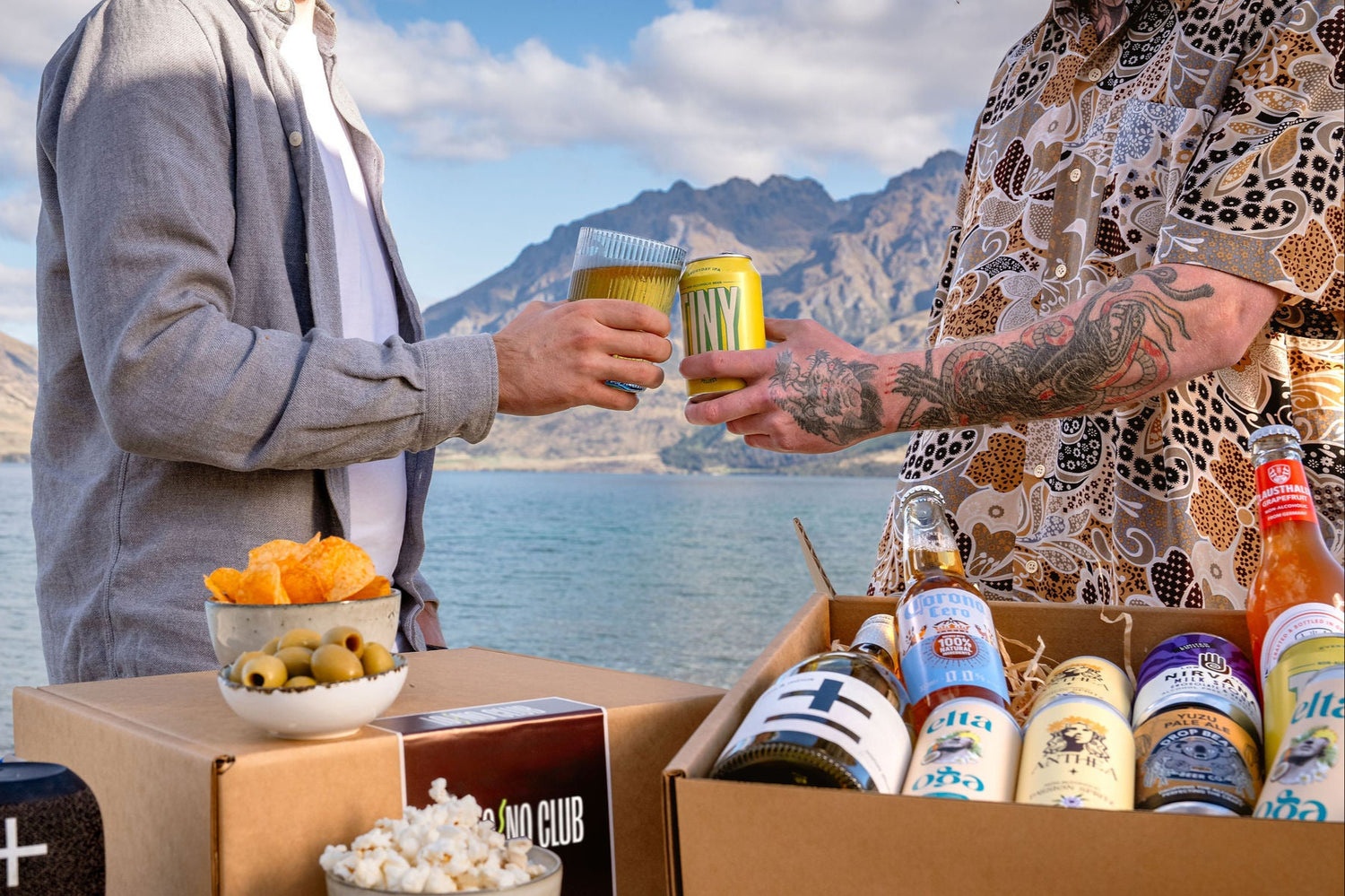 Two people cheering non alcoholic drinks in-front of Lake Wakatipu with two LONO subscription boxes in front of them.